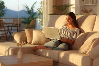 Mutlu kadın gün batımında kanepeye oturmuş kişisel bilgisayar kullanıyor.