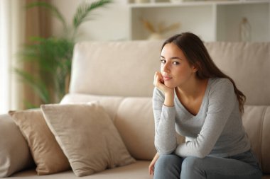 Evde rahat bir kadın kanepeye oturmuş kameraya bakıyor.