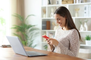 Evdeki bir odada dizüstü bilgisayarın yanında telefonu kullanan mutlu kadın.