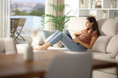 Evdeki rahat koltukta oturan bir kadın dizüstü bilgisayar kullanıyor.