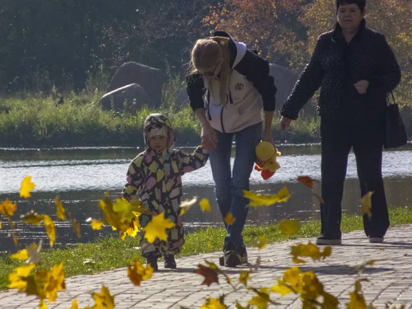 Γυναίκα με παιδί. Φθινοπωρινός χρόνος, φθινόπωρο στην πόλη, αστικό φθινοπωρινό τοπίο με λιμνούλα, άνθρωποι που περπατούν και άλλη διασκέδαση. Τα φύλλα πετούν και γυρίζουν — Φωτογραφία Αρχείου