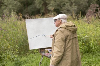 Yaşlı bir adam yağlı boyalarla resim yapmakla ilgileniyor. Beyaz bir karton üzerine doğal bir manzaranın yağlı resimlerini çizmek..