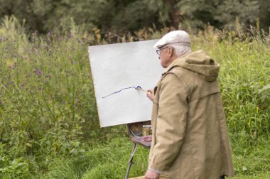 Yaşlı bir adam yağlı boyalarla resim yapmakla ilgileniyor. Beyaz bir karton üzerine doğal bir manzaranın yağlı resimlerini çizmek..