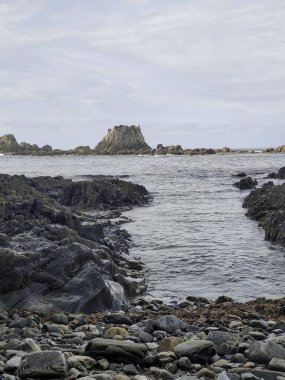 İspanya, Asteria 'daki kayalık bir sahil şeridinde sakin bir manzara. Karanlık kayalar ve çakıl taşları bulutlu bir gökyüzünün altında sakin okyanus suyuyla buluşuyor.