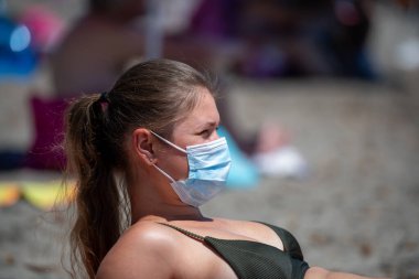 Plajda dinlenen genç bir kadın COVID-19 yüzünden koruyucu yüz maskesi takıyor.