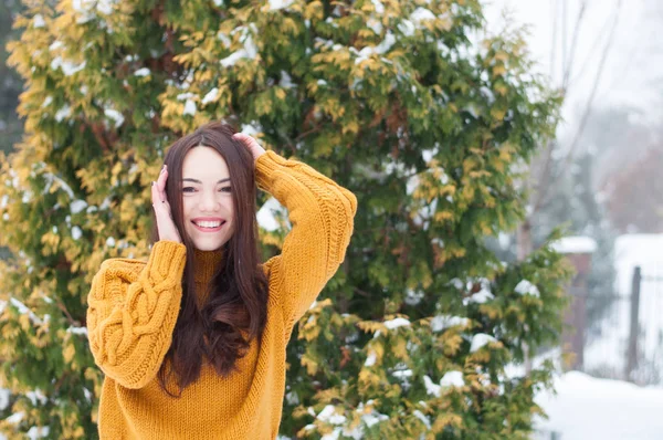 Portrait of young dark haired beautiful smiling woman in yellow knitted sweater and blue jeans on snow covered garden background. Winter snowy landscape
