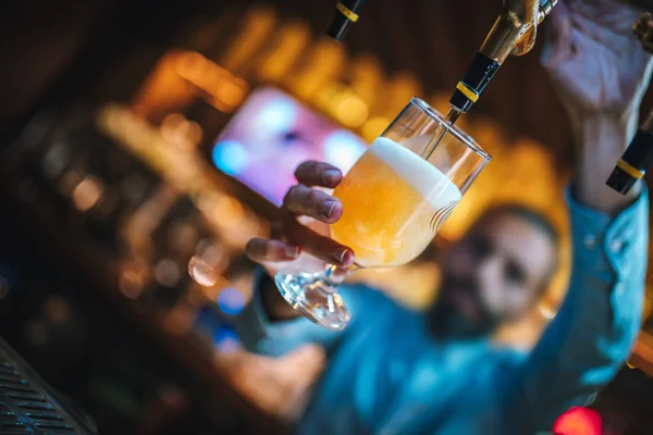 Barman, brewer or bartender filling glass with beer in the bar. Barman is pouring lager beer to glass from beer taps. Bar or night club interior