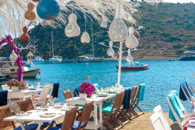 Türkiye'nin Bodrum kenti Gumusluk'ta balık restoranı veya kafe ve begonviller çiçeği manzarası. Güzel Ege Denizi'ne yakın Bodrum ilçesinde Ege sahil tarzı renkli sandalyeler, masalar ve çiçekler.
