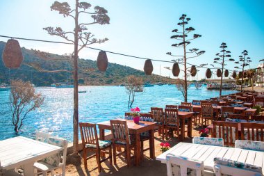 Türkiye'nin Bodrum kenti Gumusluk'ta Bodrum sahilinde balık restoranı veya kafe ve begonviller çiçeği manzarası. Güzel Ege Denizi'ne yakın Bodrum ilçesinde Ege sahil tarzı renkli sandalyeler, evler, marina, yelkenli tekneler, yatlar, masalar ve çiçekler.