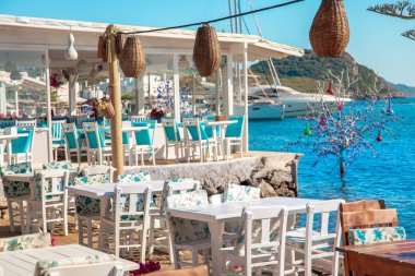 Türkiye'nin Bodrum kenti Gumusluk'ta Bodrum sahilinde balık restoranı veya kafe ve begonviller çiçeği manzarası. Güzel Ege Denizi'ne yakın Bodrum ilçesinde Ege sahil tarzı renkli sandalyeler, evler, marina, yelkenli tekneler, yatlar, masalar ve çiçekler.