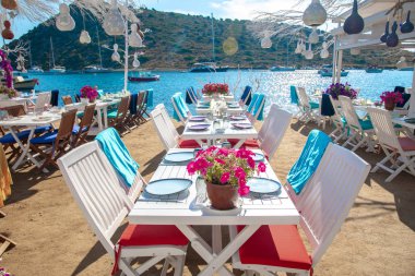 Türkiye'nin Bodrum kenti Gumusluk'ta Bodrum sahilinde balık restoranı veya kafe ve begonviller çiçeği manzarası. Güzel Ege Denizi'ne yakın Bodrum ilçesinde Ege sahil tarzı renkli sandalyeler, evler, marina, yelkenli tekneler, yatlar, masalar ve çiçekler.