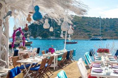 Türkiye'nin Bodrum kenti Gumusluk'ta Bodrum sahilinde balık restoranı veya kafe ve begonviller çiçeği manzarası. Güzel Ege Denizi'ne yakın Bodrum ilçesinde Ege sahil tarzı renkli sandalyeler, evler, marina, yelkenli tekneler, yatlar, masalar ve çiçekler.