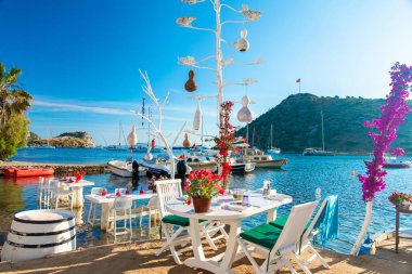 Türkiye'nin Bodrum kenti Gumusluk'ta Bodrum sahilinde balık restoranı veya kafe ve begonviller çiçeği manzarası. Güzel Ege Denizi'ne yakın Bodrum ilçesinde Ege sahil tarzı renkli sandalyeler, evler, marina, yelkenli tekneler, yatlar, masalar ve çiçekler.