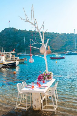 Türkiye'nin Bodrum kenti Gumusluk'ta Bodrum sahilinde balık restoranı veya kafe ve begonviller çiçeği manzarası. Güzel Ege Denizi'ne yakın Bodrum ilçesinde Ege sahil tarzı renkli sandalyeler, evler, marina, yelkenli tekneler, yatlar, masalar ve çiçekler.