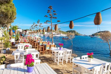 Türkiye'nin Bodrum kenti Gumusluk'ta Bodrum sahilinde balık restoranı veya kafe ve begonviller çiçeği manzarası. Güzel Ege Denizi'ne yakın Bodrum ilçesinde Ege sahil tarzı renkli sandalyeler, evler, marina, yelkenli tekneler, yatlar, masalar ve çiçekler.