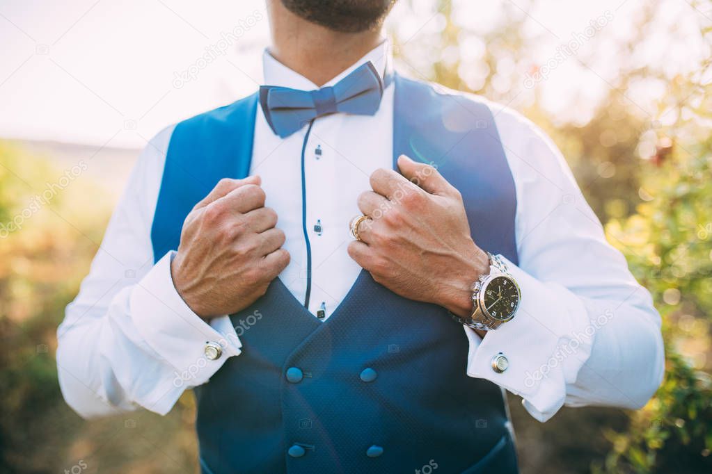 Groom hands holding and adjusting blue vest. Classy, elegant groom posing with blue bowtie, watch, vest and white shirt