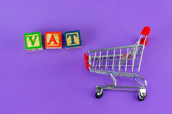 VAT (Value-Added Tax) on household goods concept. Trolley with alphabet (V.A.T) block on purple background.