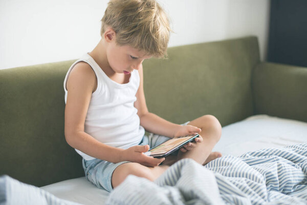Boy fair hair playing tablet games laying bed. home interior. modern device technologies Selective focus