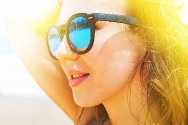 Deniz kıyısı üzerinde mavi güneş gözlüğü portre rahatlatıcı genç siyah dalgalı saçlı kadın kapatmak kirişler güneş güneşli gün içinde Sunbathes. Tropikal iklim dinlenme tatil kavramı güneş ışığı etkisi zevk deniz kıyısında ayakta