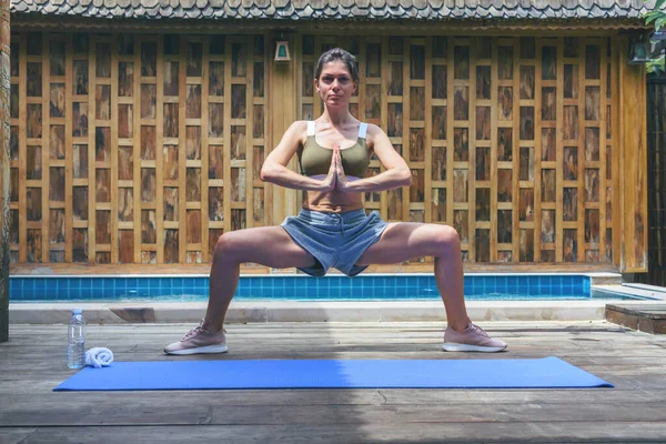 Genç, formda bir kadın, ahşap arka planda, yüzme havuzunun yanında yoga esneme egzersizi yapıyor. Kadın havuz güvertesinde meditasyon yapıyor. Kadın yoga pozları veriyor. Nefes alıştırması..