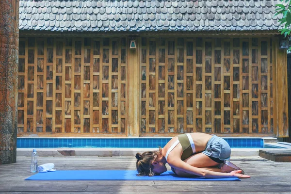 Genç, formda bir kadın çocuk pozu veriyor. Yoga esneme egzersizi, ahşap arka planda yüzme havuzunun yanında. Kadın havuz güvertesinde meditasyon yapıyor. Nefes alıştırması..