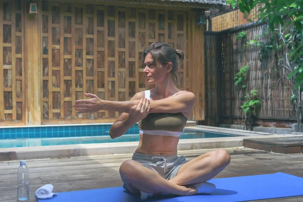 Genç, formda bir kadın, ahşap arka planda, açık havada, yüzme havuzunun yanında kol egzersizi yapıyor. Kadın yoga pozu veriyor. Dışarıda dinleniyor.