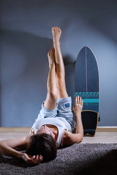 Girl lying on the floor with legs up the wall is imitating sitting near longboard and waiting the end of quarantine. legs upside down. Outdoor activities. Sport. Background. Wall paper. Break. Rest.