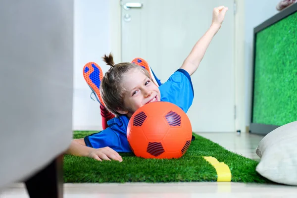 サッカーボールを持つ少年が横になり 隔離中に自宅のテレビ画面の近くの緑のフィールドに手を上げました 家にいて スポーツ活動 コロナウイルスの状況 サッカーゲーム屋内 — ストック写真