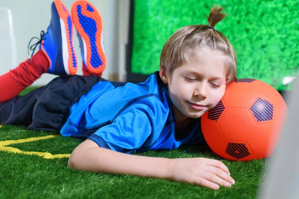 Young football player tired to play at home on green field near TV screen during the quarantine. Stay at home. Sport activities. Coronavirus situation. Sleeping boy. Kids activities.