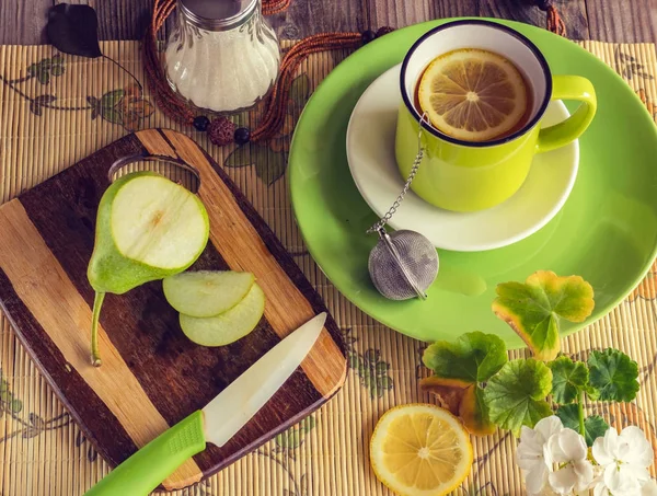 Healthy breakfast. Tea with lemon in a green cup. Near the sugar bowl, a board with a green pear and a knife. Atmospheric photo. Fresh bright colors. Top view.