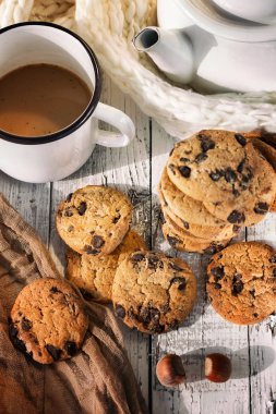 Beyaz bardakta sütlü kahve. Beyaz bir çaydanlığın yanında ve çikolatalı kurabiyeler.