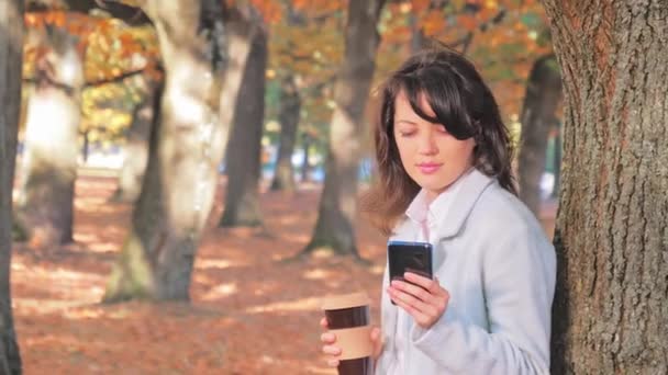 Jonge vrouw drinkt koffie en maakt gebruik van smartphone in de herfst park, kleurrijke bomen — Stockvideo
