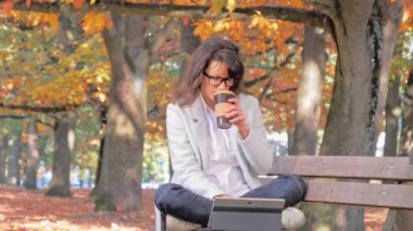 Tabletle çalışan kadın bankta oturuyor, sonbaharda parkta.