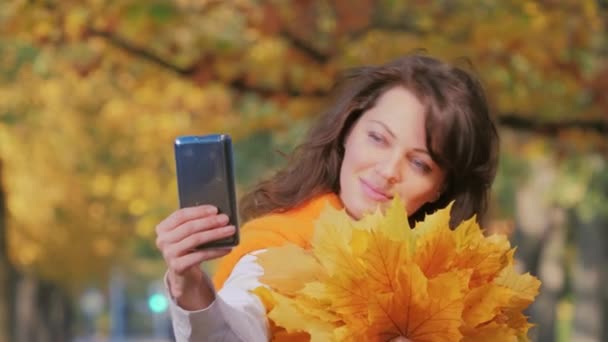 Junge lächelnde Frau mit Herbstblättern macht Selfie auf Smartphone — Stockvideo