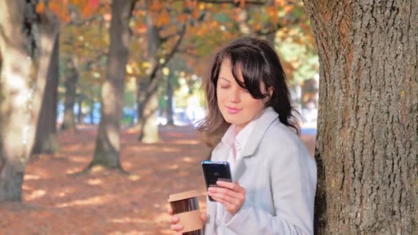 Mujer joven bebe café y utiliza teléfono inteligente en el parque de otoño . — Vídeos de Stock