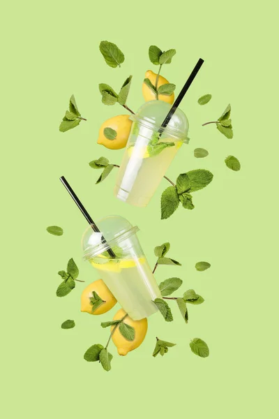 Літаючий освіжаючий лимонад з пластикових чашок з літаючими лимонами та листя м'яти на зеленому фоні. Джар повний холодного коктейлю. Свіжа концепція. Знущання . Стокове Зображення