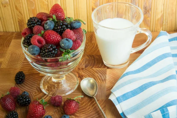 Berry raspberries strawberry blueberries blueberries in a glass vase on a wooden set of bars bars. A useful berry breakfast on a wooden table with a striped blue and white towel, a silver spoon and a glass of milk