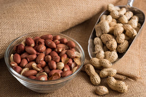 Glass bowl and metal scoop full of peeled peanuts and peanuts in nutshell on sackcloth