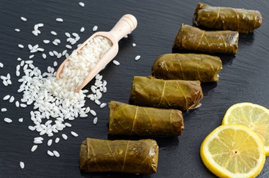 Limon dilimleri ve çiğ pirinç dolu minyatür ahşap kepçe ile siyah çanak üzerinde lezzetli doldurulmuş üzüm yaprakları (Akdeniz mutfağının geleneksel dolma)