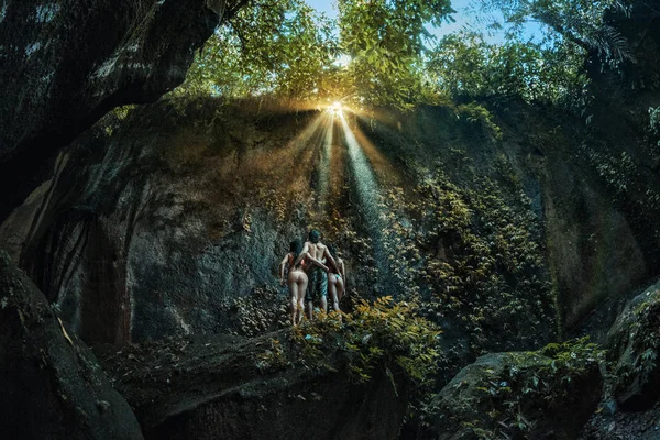 Three persons in cave, two girls and one man standing on big stone inside a cave, looking at sunlight falling through threes, view from back and below