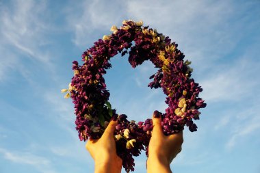 Wreath of Pink Flowers on Blue Sky Background. Female hands holding a Crown of purple flowers on a background of blue sky with white clouds. Hands with a wreath of Midsummer flowers                         clipart