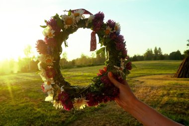 Vahşi çiçeklerden oluşan bir çelenk ve akşam güneşinin arka planında işaretli kırmızı ve beyaz bir kurdele tutan kadın eller.                               