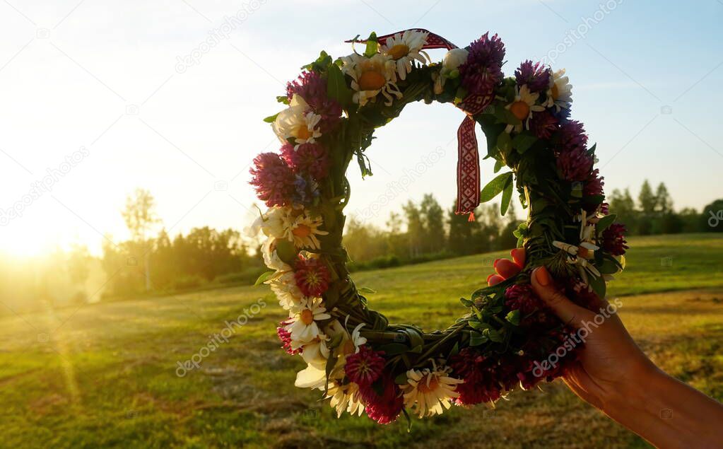 Female hands holding a wreath of wild flowers and a red and white ribbon with signs on a background of evening sunset                               