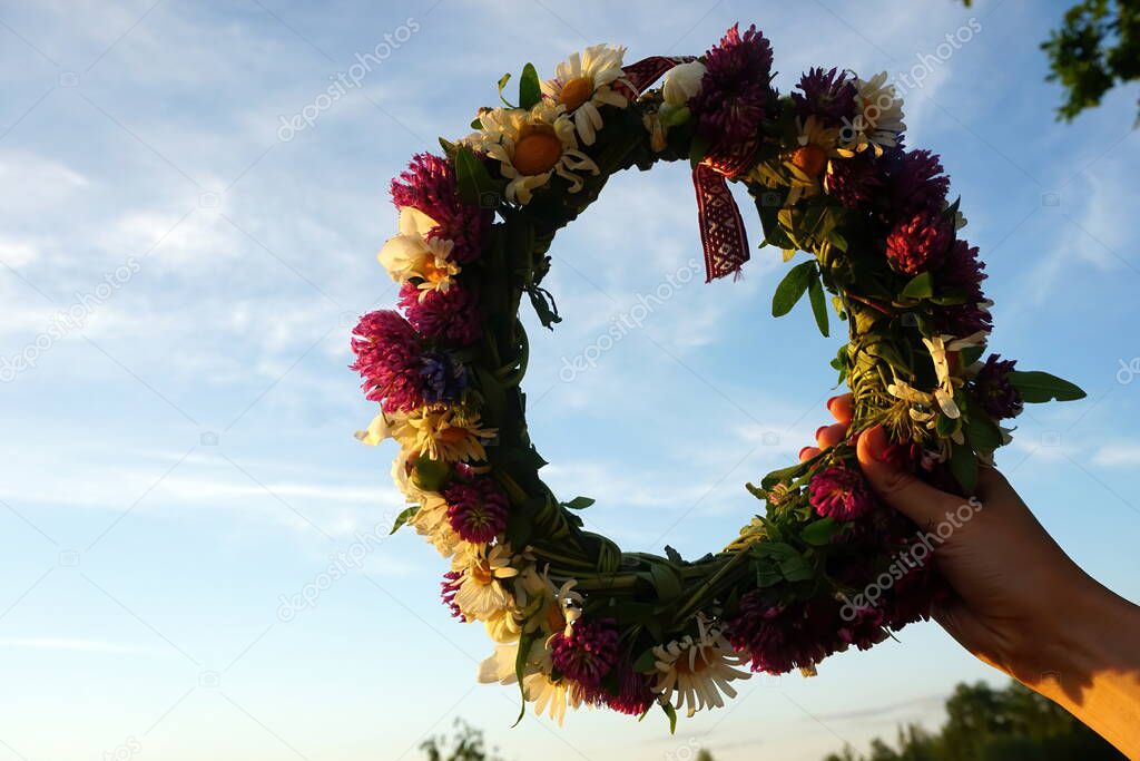 Female hands holding a wreath of wild flowers and a red and white ribbon with signs on a background of blue sky with white clouds              