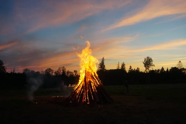 A large bonfire burns at midsummer night, with the sun setting in the background