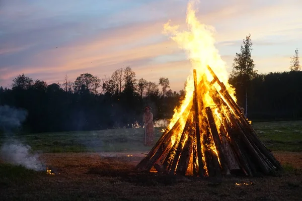 A large bonfire burns at midsummer night, with the sun setting in the background