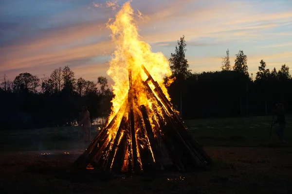 A large bonfire burns at midsummer night, with the sun setting in the background