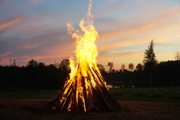 A large bonfire burns at midsummer night, with the sun setting in the background