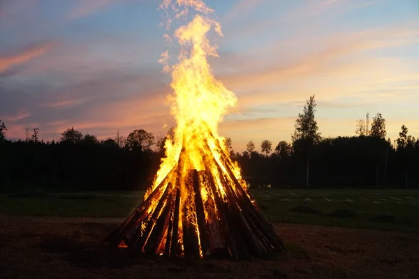 A large bonfire burns at midsummer night, with the sun setting in the background