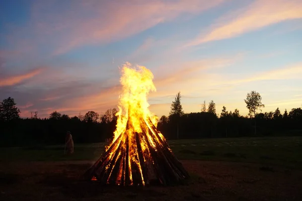 A large bonfire burns at midsummer night, with the sun setting in the background